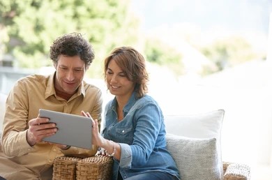A smiling couple sits on a sofa, looking at a tablet together. They seem relaxed and content, with a blurry, sunlit garden in the background.