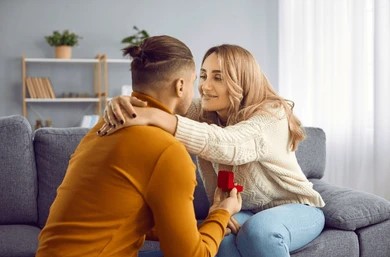 A man kneels and holds a ring box, proposing to a smiling woman on a gray couch in a cozy living room. She leans in, touching his face affectionately.