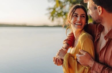 A smiling couple embraces near a tranquil lake at sunset. The woman in a yellow top smiles at the man, creating a warm, joyful atmosphere.