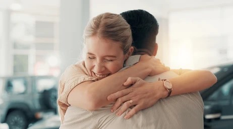 A man and woman share a warm hug in a car dealership, with cars visible in the background.
