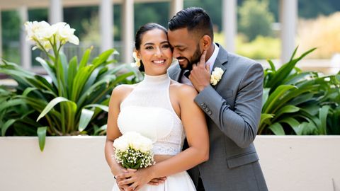 A bride in a white dress holds a bouquet of flowers while standing in front of greenery, smiling beside a groom in a gray suit.