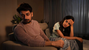 A man and woman sit on a couch looking upset and distant, with the woman holding her head and the man looking away, representing challenges in Sri Lanka matrimony and matrimony Sri Lanka. This reflects issues in marriage proposals Sri Lanka, marriage proposals in Sri Lanka, and the emotional stress couples may face when using a wedding proposal website or mangala yojana services before a Poruwa ceremony.