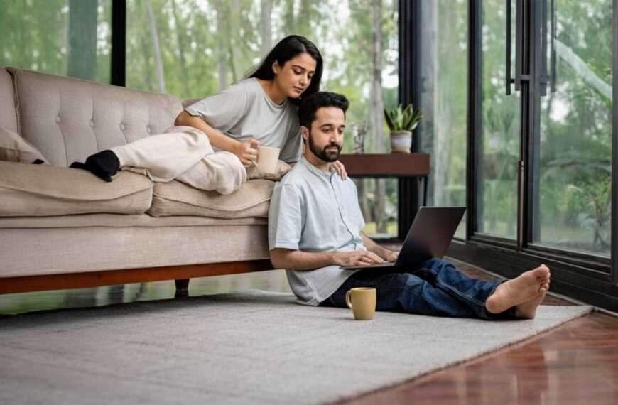 A man and woman are seated on the floor, engaged with a laptop between them.