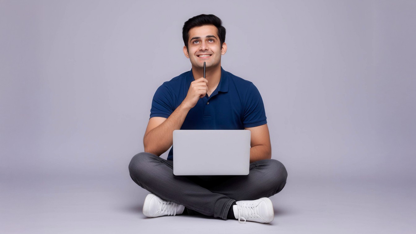 A man sits cross-legged on the floor with a laptop on his lap, smiling thoughtfully while holding a pen to his chin, representing productivity and focus in SriLanka matrimony and matrimony Sri Lanka, while exploring marriage proposals Sri Lanka and planning a traditional Poruwa ceremony.