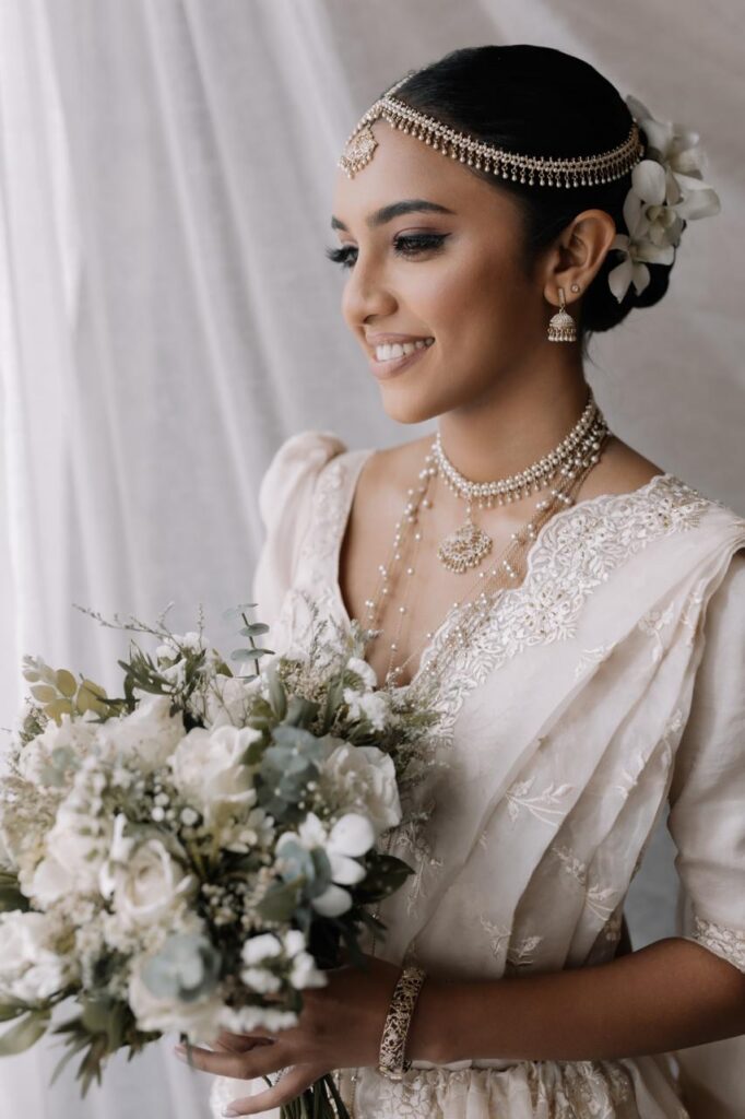 A smiling bride in an elegant white dress holding a lush bouquet of white and green flowers, wearing intricate jewelry and floral hair accessories, representing Sri Lankan matrimony, matrimony Sri Lanka, marriage proposal Sri Lanka, marriage proposals in Sri Lanka, marriage proposals Sri Lanka, mangala yojana, wedding proposal website services, and traditional bridal beauty through Poruwa.lk | ශ්රී ලාංකීය මංගල යෝජනා.