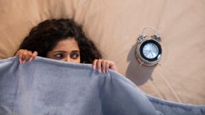 A woman with curly hair hides under a blue blanket on a bed, looking anxious, with an alarm clock beside her, reflecting stress or worry about upcoming marriage proposals in Sri Lanka, matrimony decisions, Poruwa ceremonies, and wedding proposal websites like Poruwa.lk | ශ්‍රී ලාංකීය මංගල යෝජනා.