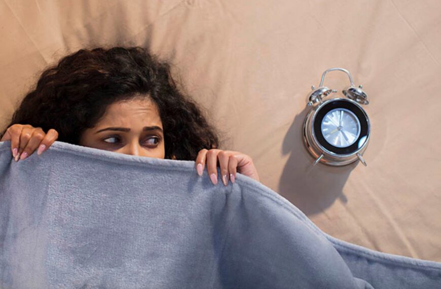 A woman with curly hair hides under a blue blanket on a bed, looking anxious, with an alarm clock beside her, reflecting stress or worry about upcoming marriage proposals in Sri Lanka, matrimony decisions, Poruwa ceremonies, and wedding proposal websites like Poruwa.lk | ශ්‍රී ලාංකීය මංගල යෝජනා.