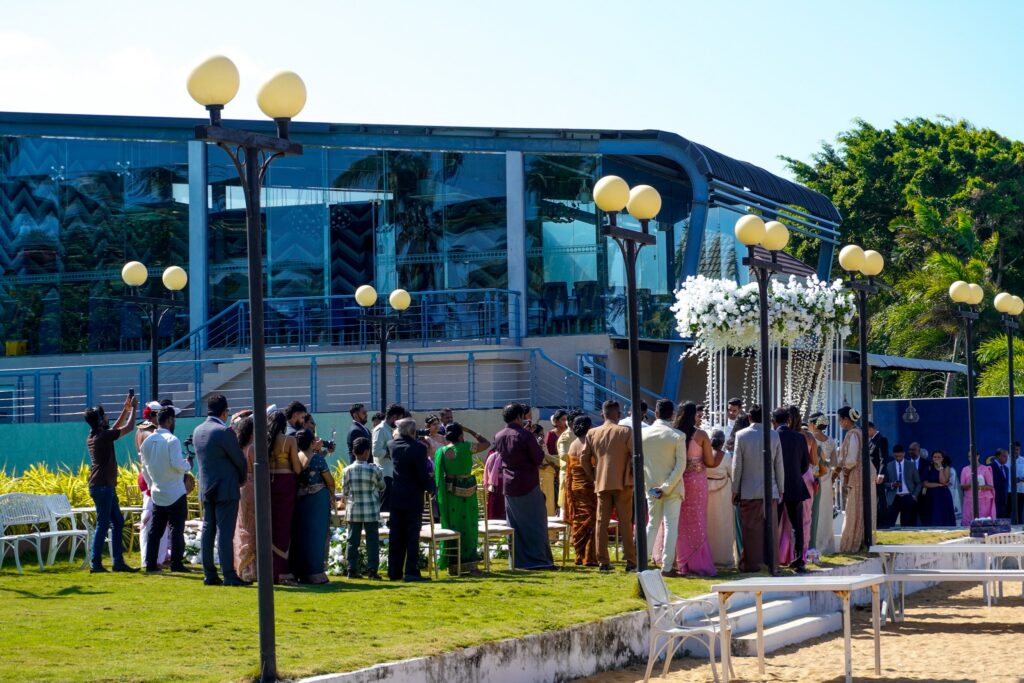 A group of people in formal attire gathered outdoors for a wedding ceremony near a modern glass building under a floral arch, representing matrimony Sri Lanka celebrations, Poruwa ceremony traditions, marriage proposals in Sri Lanka, and lanka brides and grooms events.