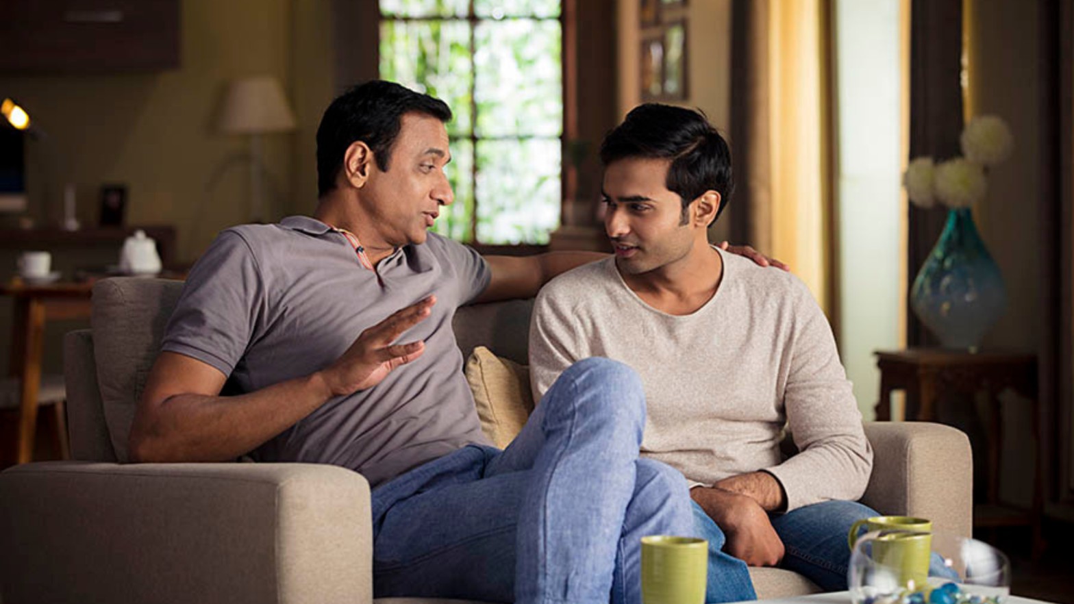 Two Sri Lankan men engaged in a thoughtful conversation while sitting on a couch in a cozy living room, discussing marriage proposals and family matters.