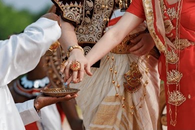 A traditional ceremony with participants in ornate attire pouring a small offering. Intricate jewelry and vibrant fabrics convey cultural richness.
