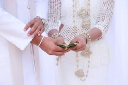 A bride and groom in traditional attire hold betel leaves together, symbolizing unity. The bride's dress is intricately designed, conveying elegance.
