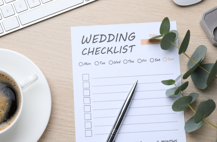 A wedding checklist on a wooden desk with a silver pen, eucalyptus leaves, a coffee cup, and a keyboard. The scene conveys organization and anticipation.