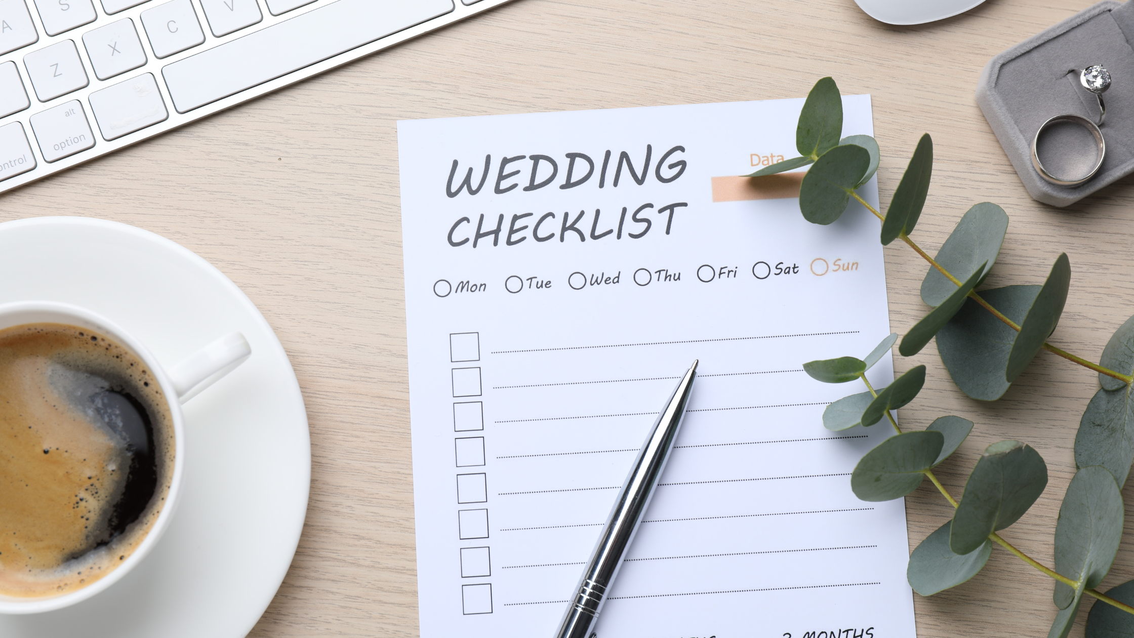A wedding checklist on a wooden desk with a silver pen, eucalyptus leaves, a coffee cup, and a keyboard. The scene conveys organization and anticipation.