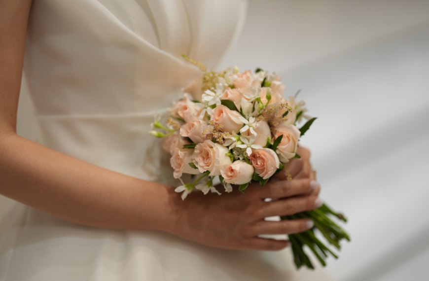 A bride in a white dress holds a bouquet of delicate pink roses and white flowers. The setting conveys elegance and a sense of serene anticipation.
