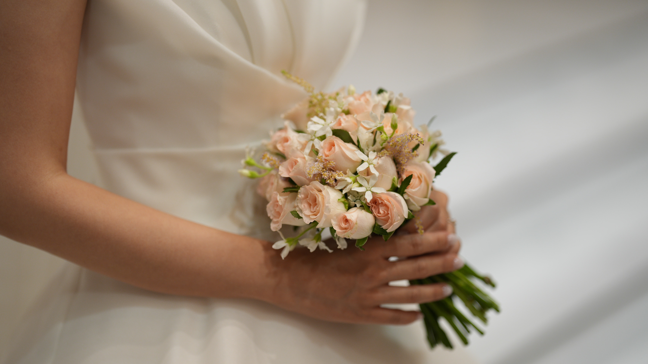 A bride in a white dress holds a bouquet of delicate pink roses and white flowers. The setting conveys elegance and a sense of serene anticipation.