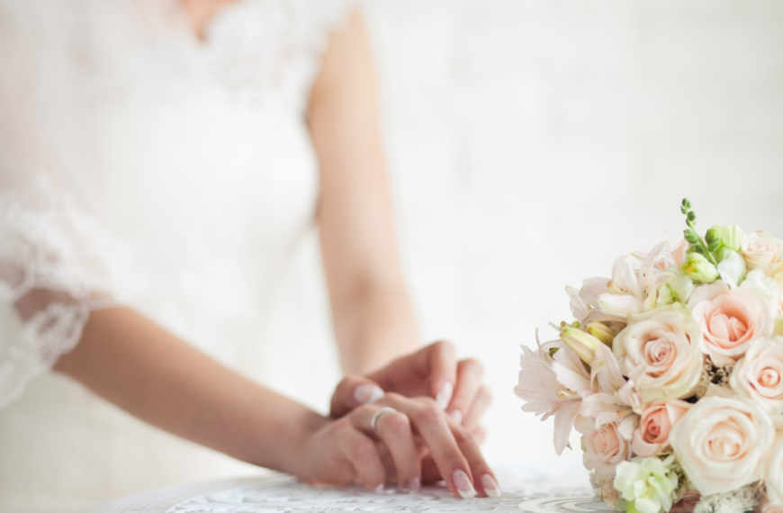 A bride in a white lace dress stands softly focused, hands resting on a lace tablecloth beside a bouquet of pale pink roses, conveying elegance and anticipation.