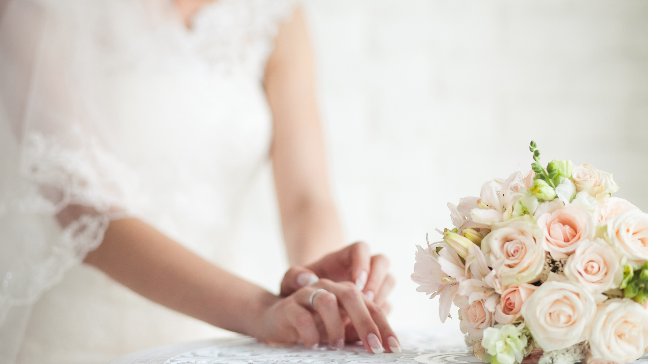 A bride in a white lace dress stands softly focused, hands resting on a lace tablecloth beside a bouquet of pale pink roses, conveying elegance and anticipation.