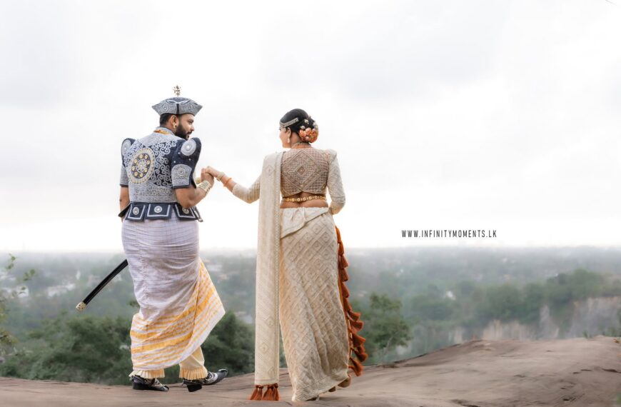 A traditionally dressed Sri Lankan wedding couple holds hands on a scenic rocky overlook, surrounded by misty green hills, capturing the romance and beauty of a Poruwa ceremony and Sri Lankan wedding celebration.