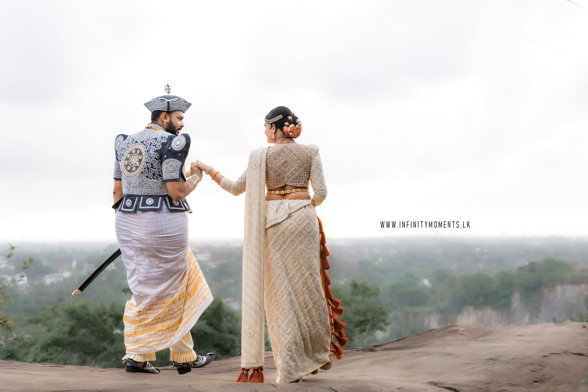 A traditionally dressed Sri Lankan wedding couple holds hands on a scenic rocky overlook, surrounded by misty green hills, capturing the romance and beauty of a Poruwa ceremony and Sri Lankan wedding celebration.