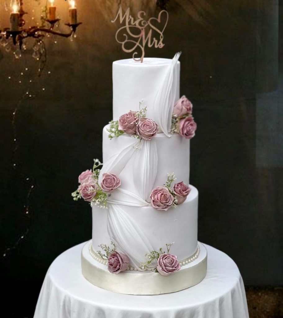 Three-tiered wedding cake adorned with delicate pink roses on each layer, representing elegant Sri Lankan wedding celebrations, mangala yojana, marriage proposals in Sri Lanka, matrimony Sri Lanka, srilanka matrimony, and special moments for Sri Lankan brides and grooms.